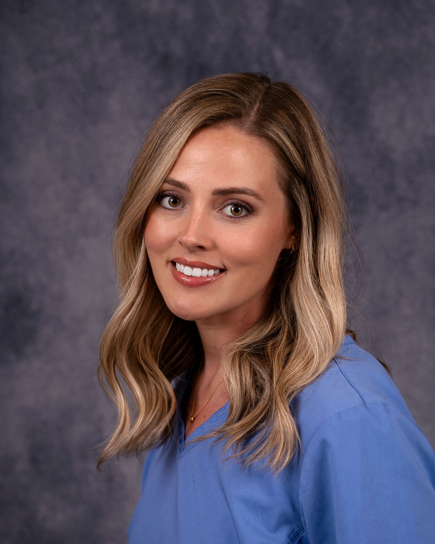 Woman with wavy blonde hair posing with friendly smile wearing light blue business attire against dark studio backdrop.