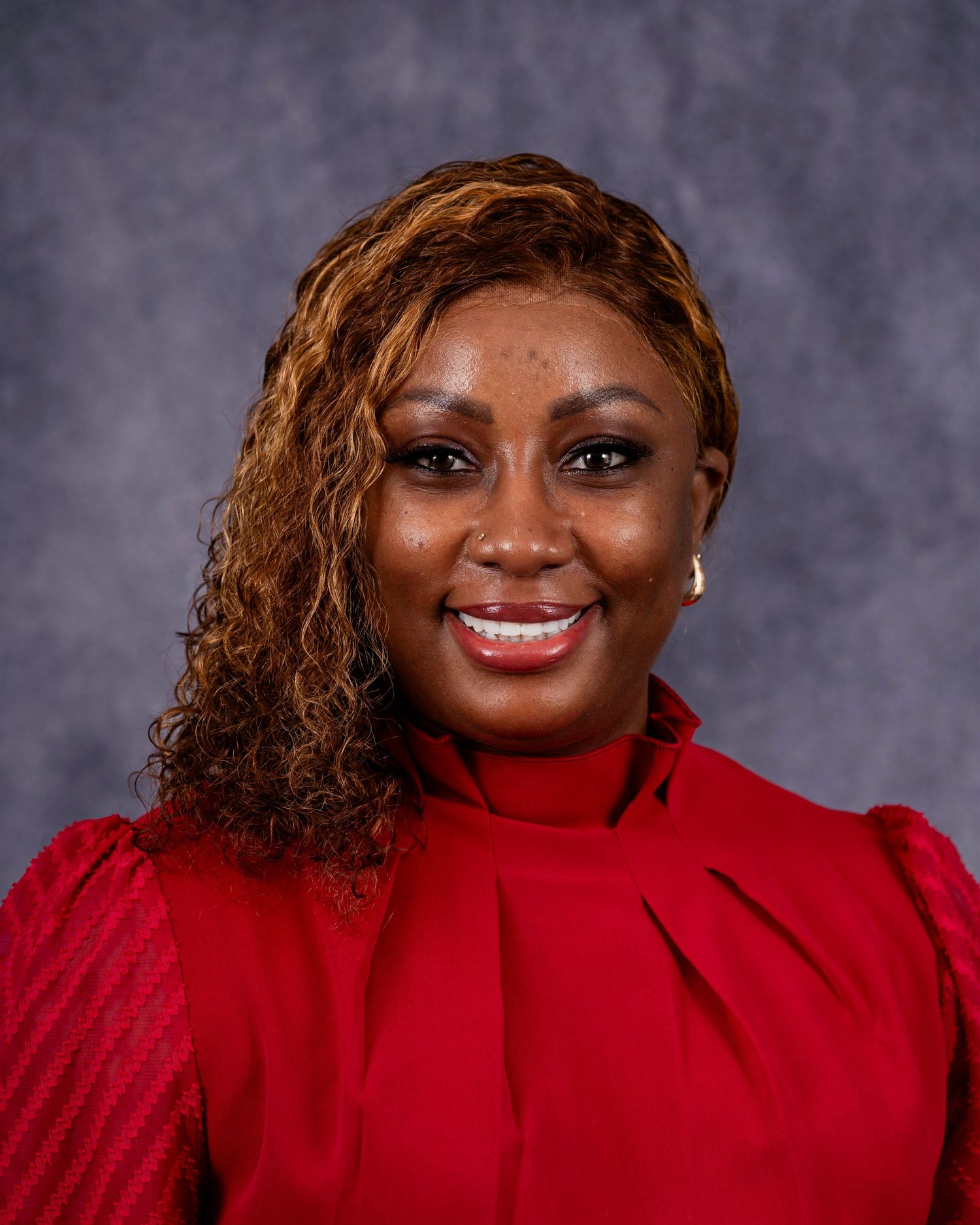 Professional headshot portrait of someone in a red sweater with curly brown hair against a gray background.