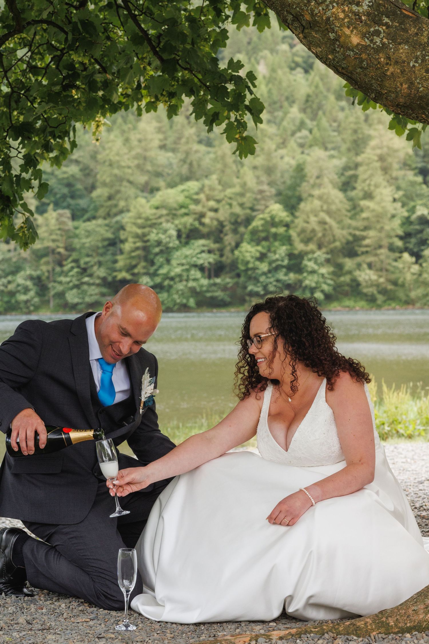 A couple shares a champagne moment by a scenic lake surrounded by green forest.