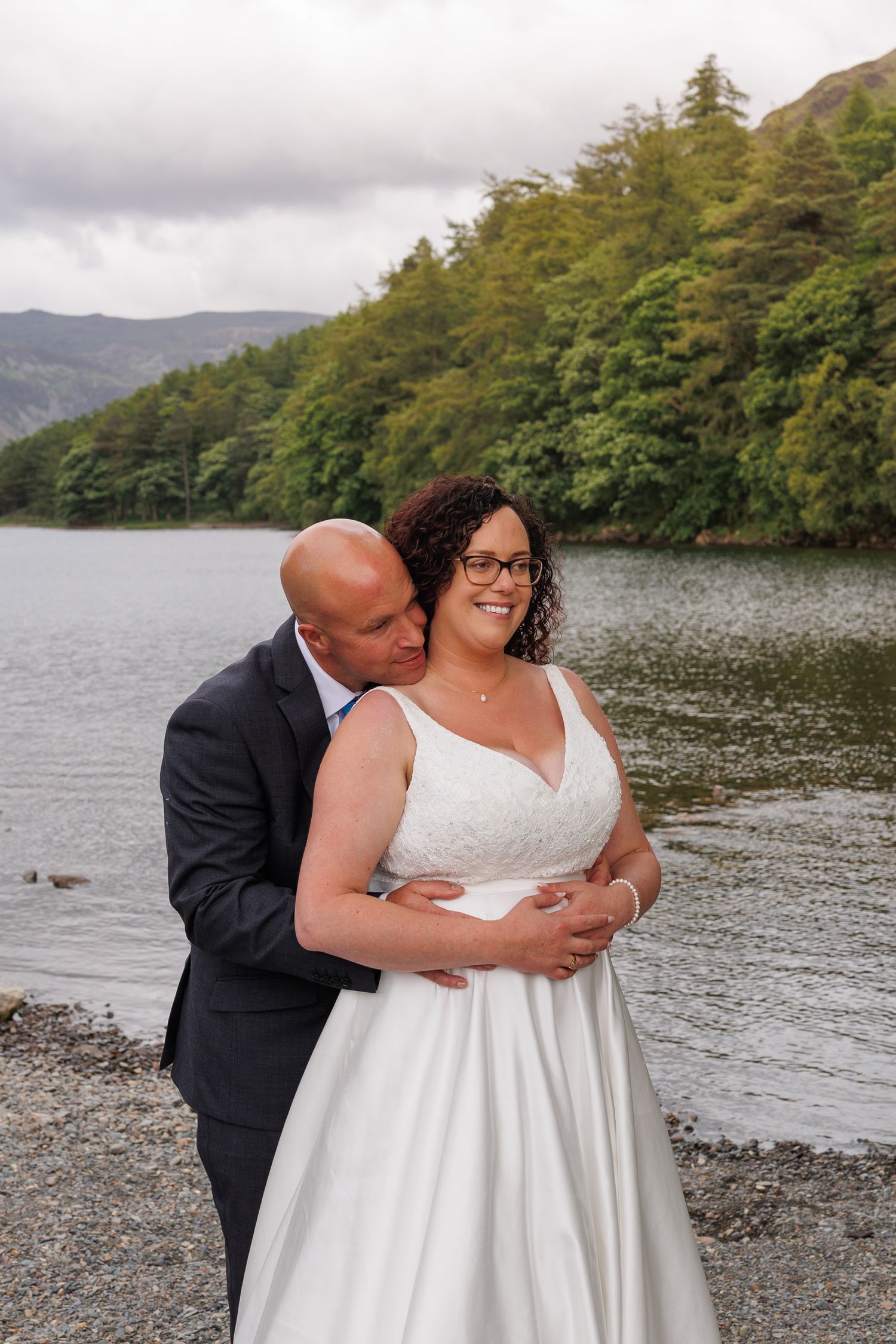 A romantic newlywed couple embraces beside a tranquil lake with mountains and forests in the background.