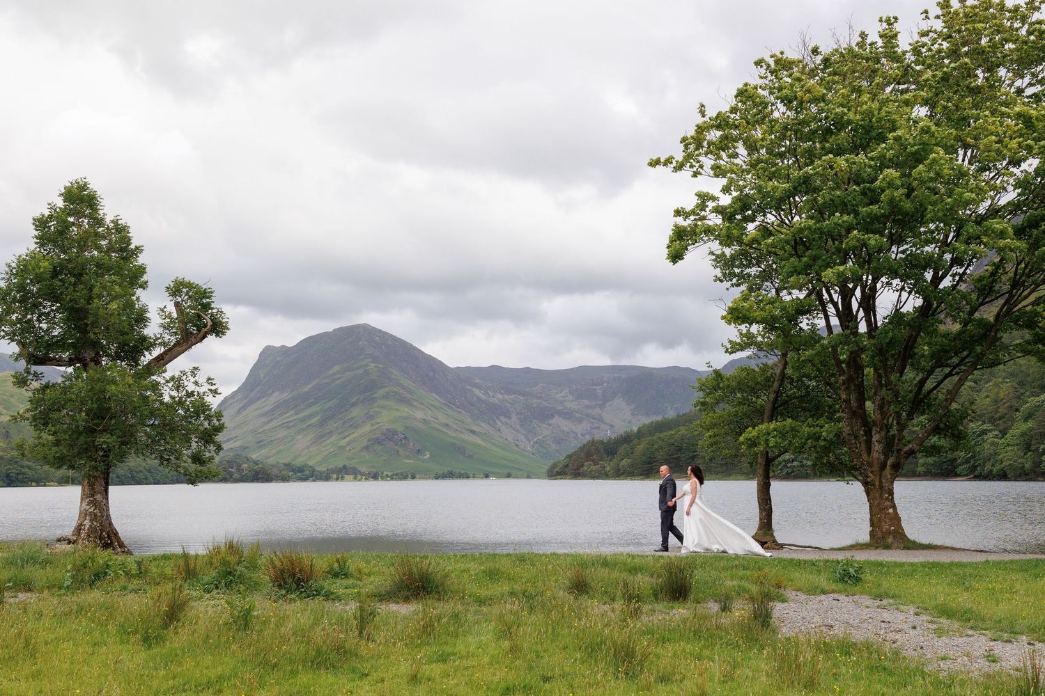 Two silhouettes in distance walk on a lovely lakeside path surrounded by trees with mountains in background.