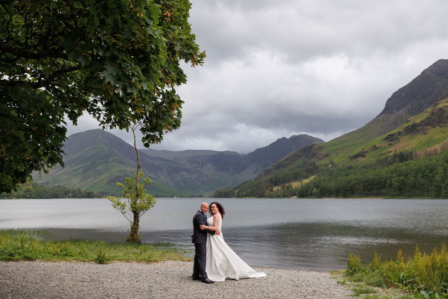 Couple in wedding attire embrace by a serene mountain lake in Scotland with dramatic cloudy skies above.