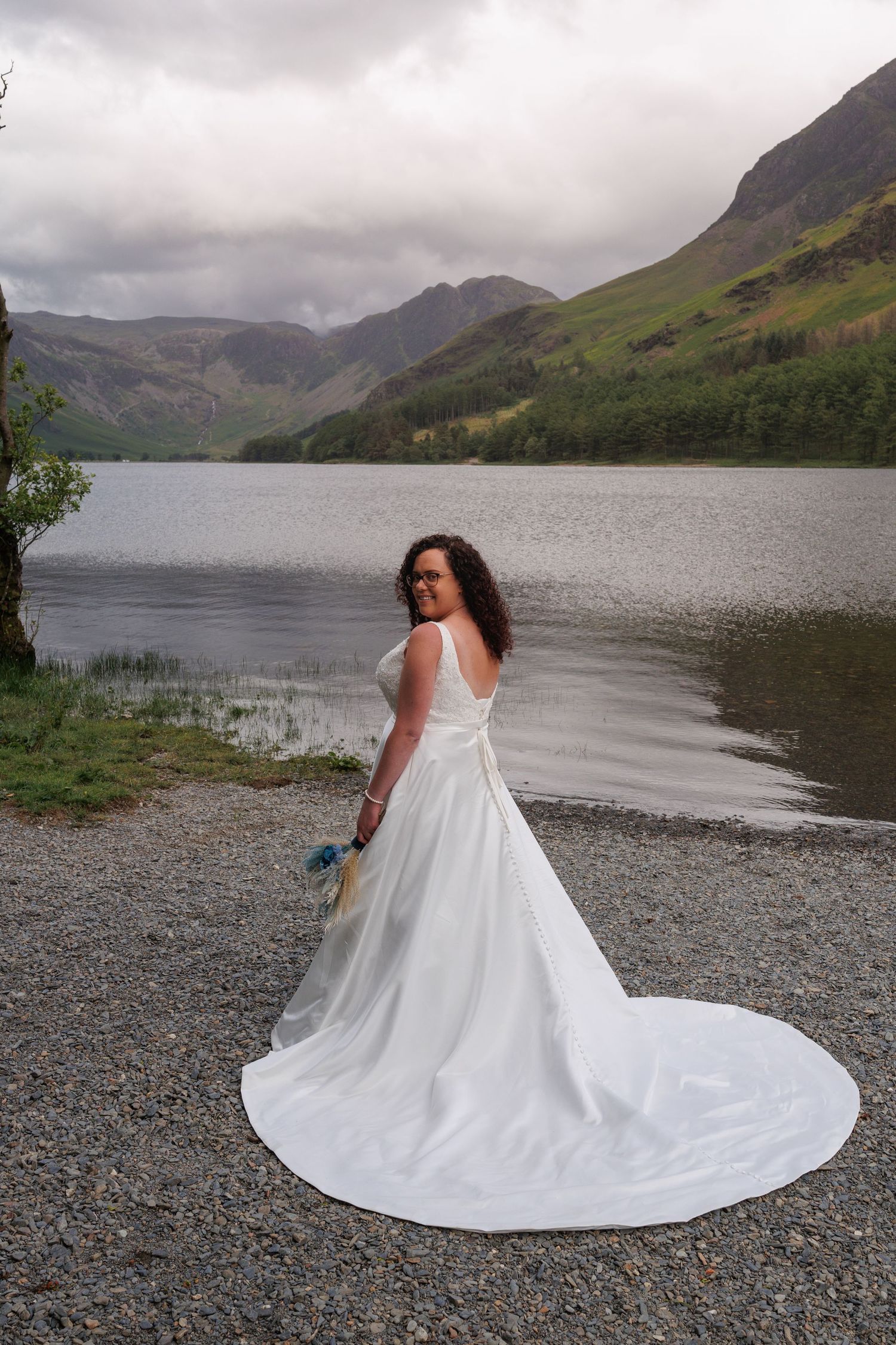 White wedding dress train flowing gracefully against rocky shoreline with stunning mountain lake backdrop.