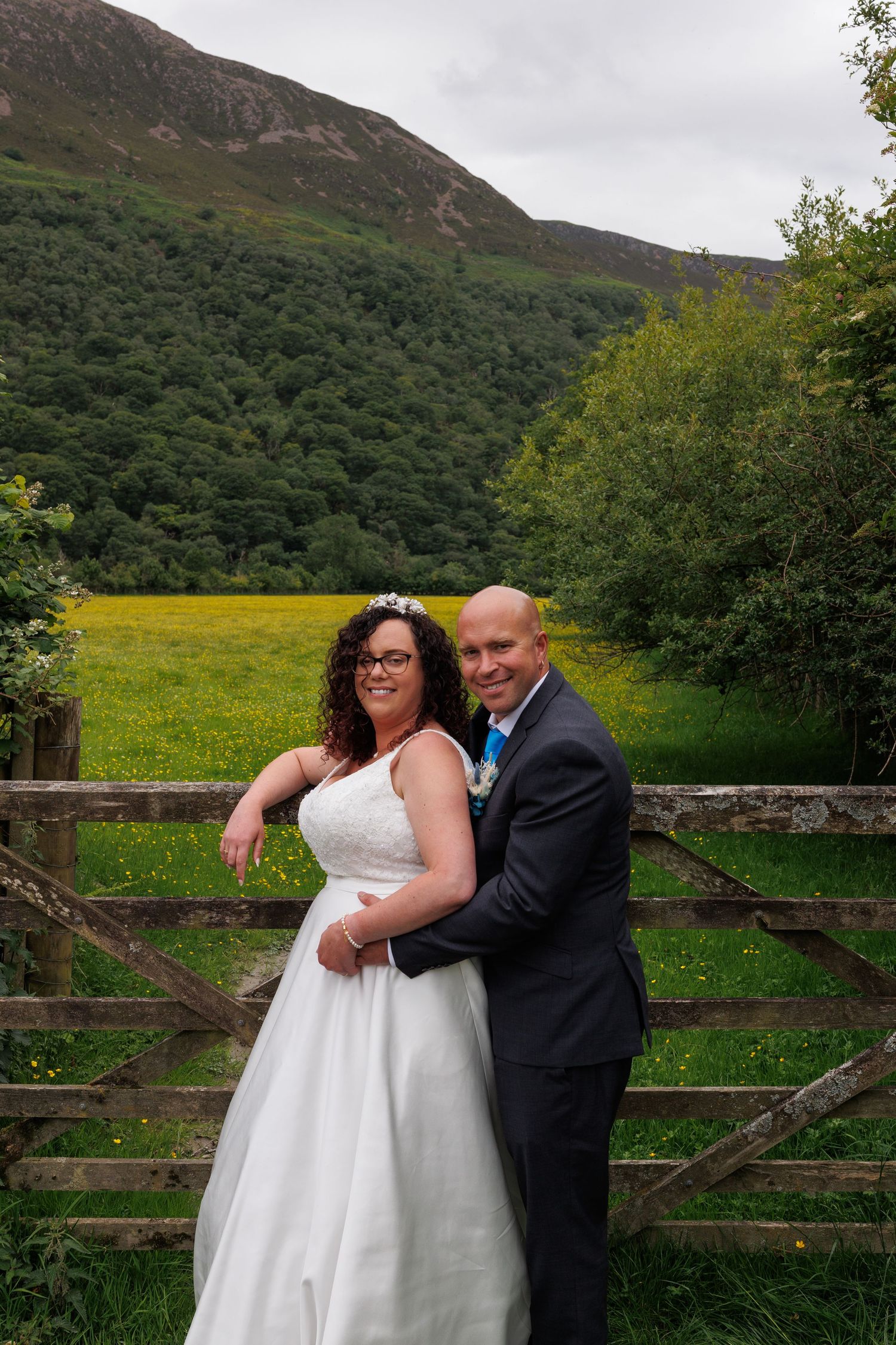 Wedding couple embracing in front of rustic wooden fence with rolling hills and mountains in background.