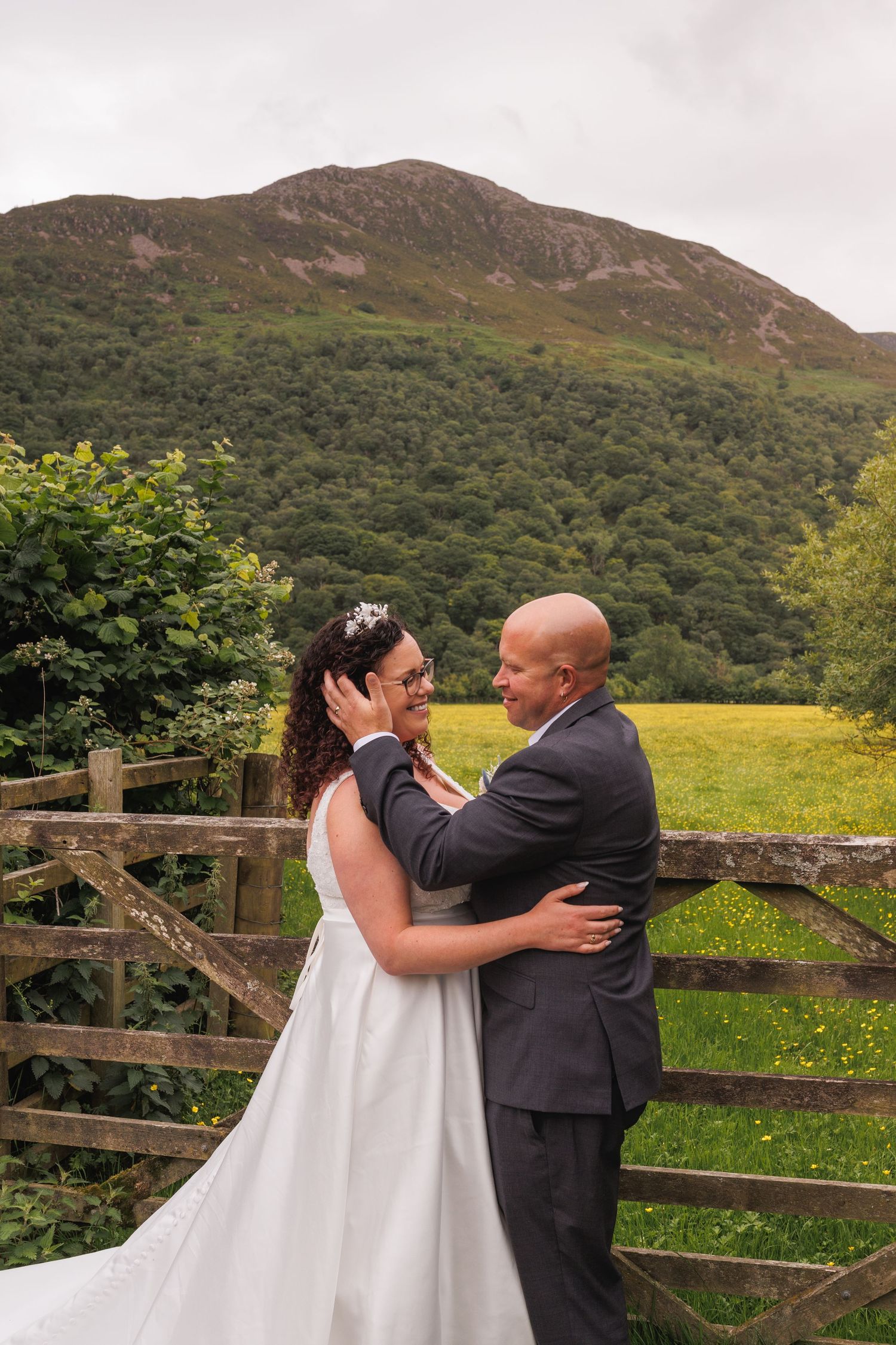 A couple shares their wedding day embrace in front of a rustic wooden fence with a scenic mountain landscape in Scotland.
