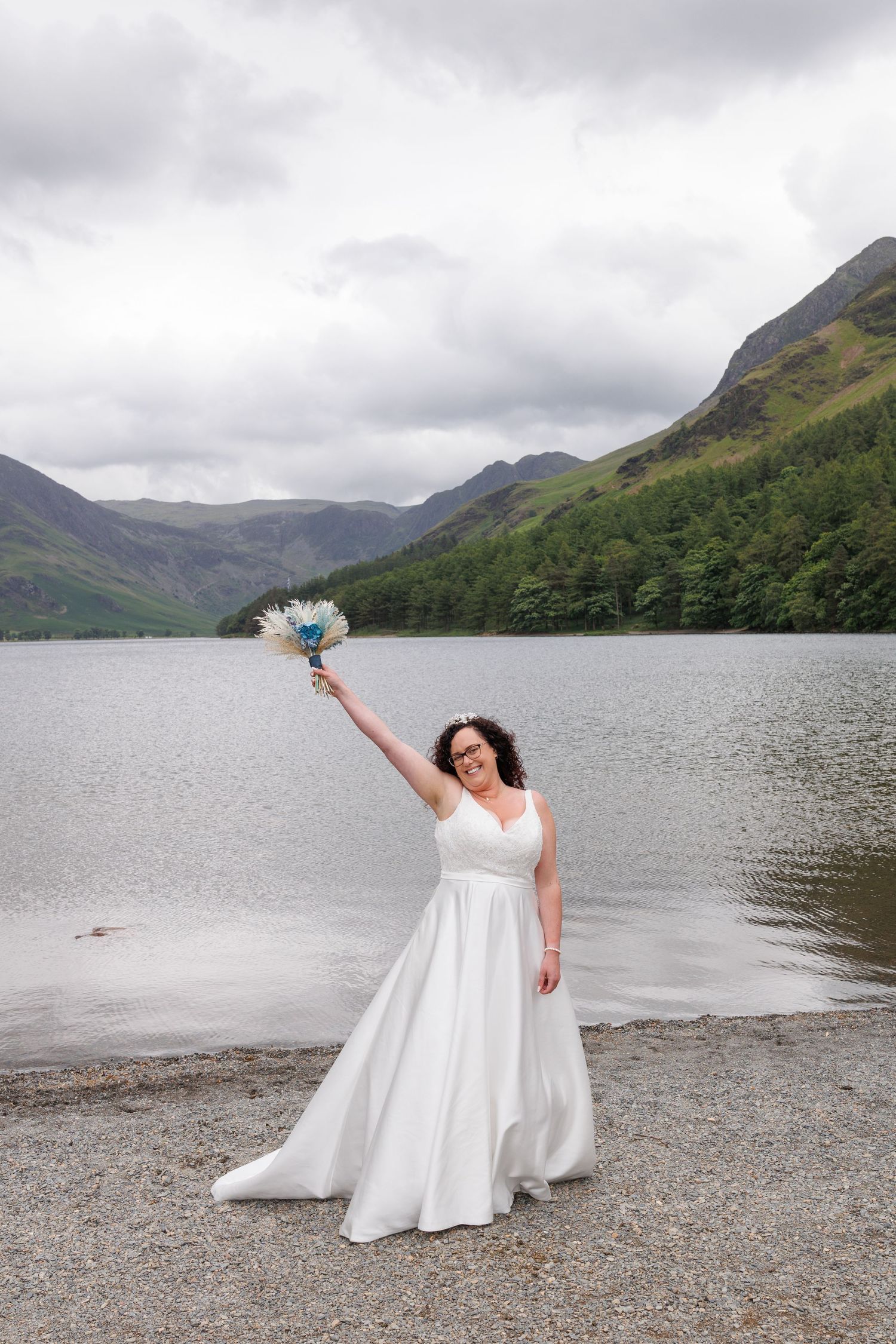 Someone in a white wedding dress poses elegantly along a scenic lakeshore with mountains in the background on a cloudy day.