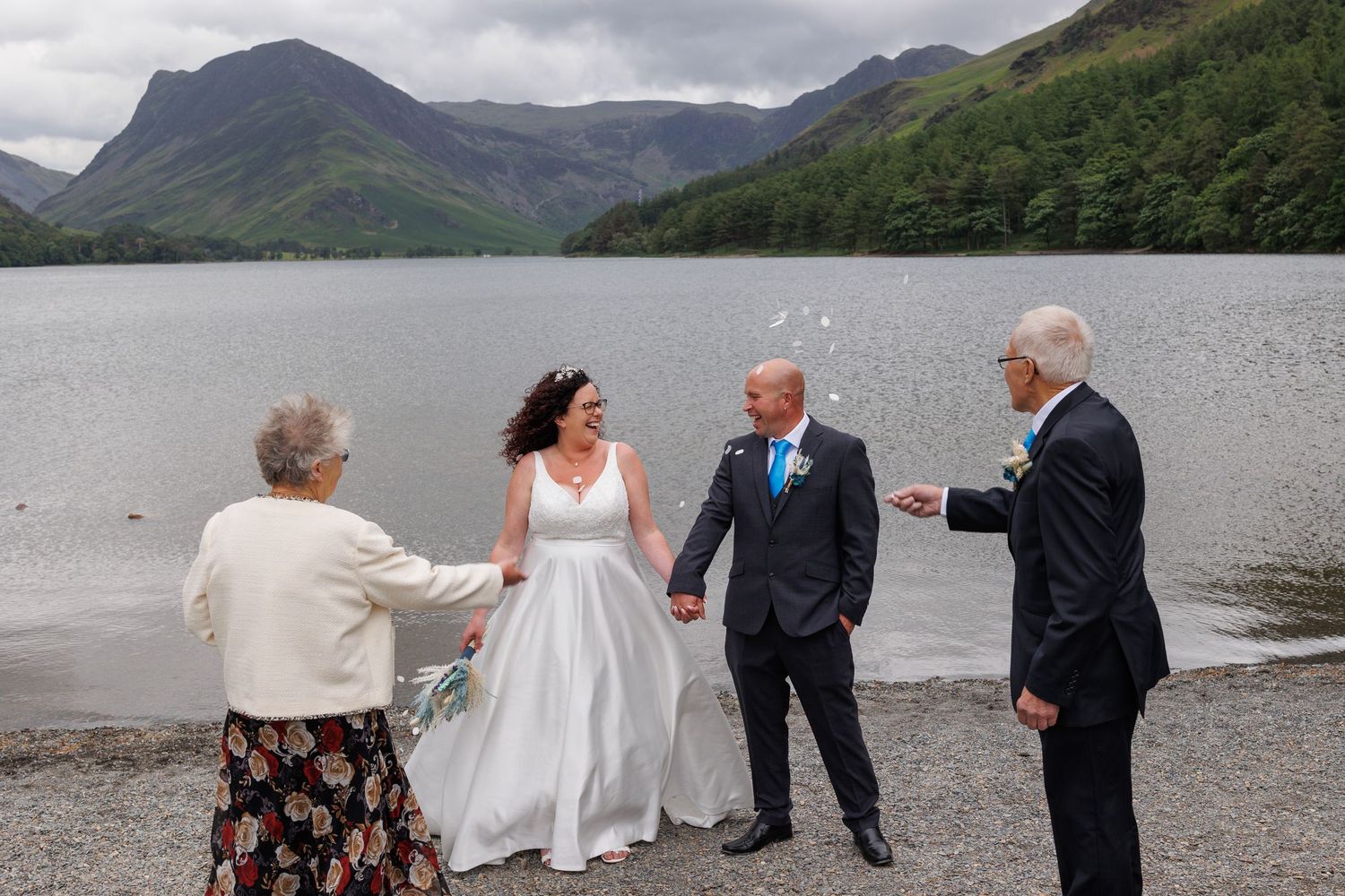 Wedding party celebrating on a scenic lakeshore with mountains and dramatic clouds in the background.
