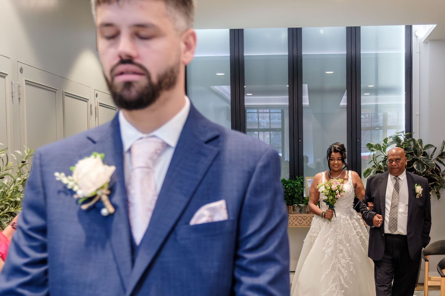 A wedding scene showing the first look moment with a blurred groom in blue suit as the bride approaches in white dress.