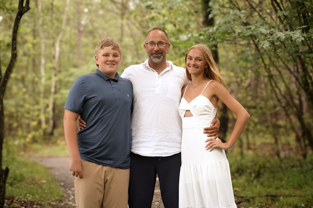 Three people pose together on a wooded path, with one wearing a white dress and others in casual attire.