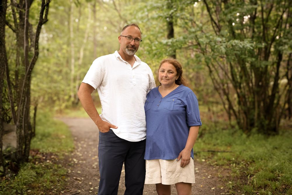 Two people stand together smiling on a wooded nature trail surrounded by green trees.
