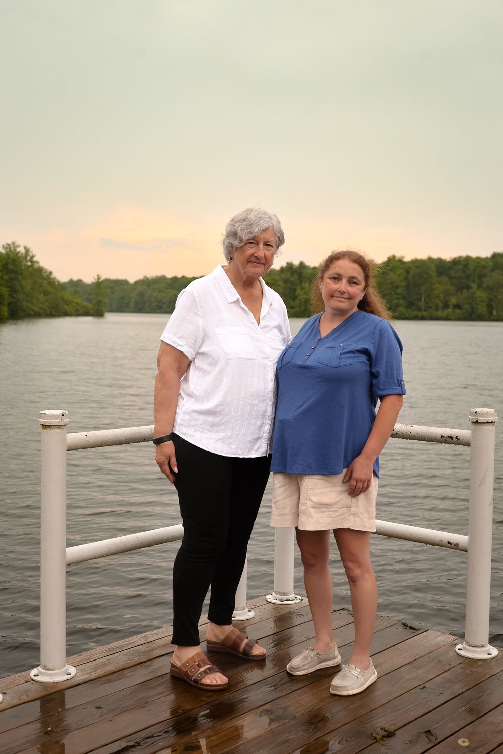 Two people stand together on a wooden dock by a serene lake at sunset, with trees visible in the background.