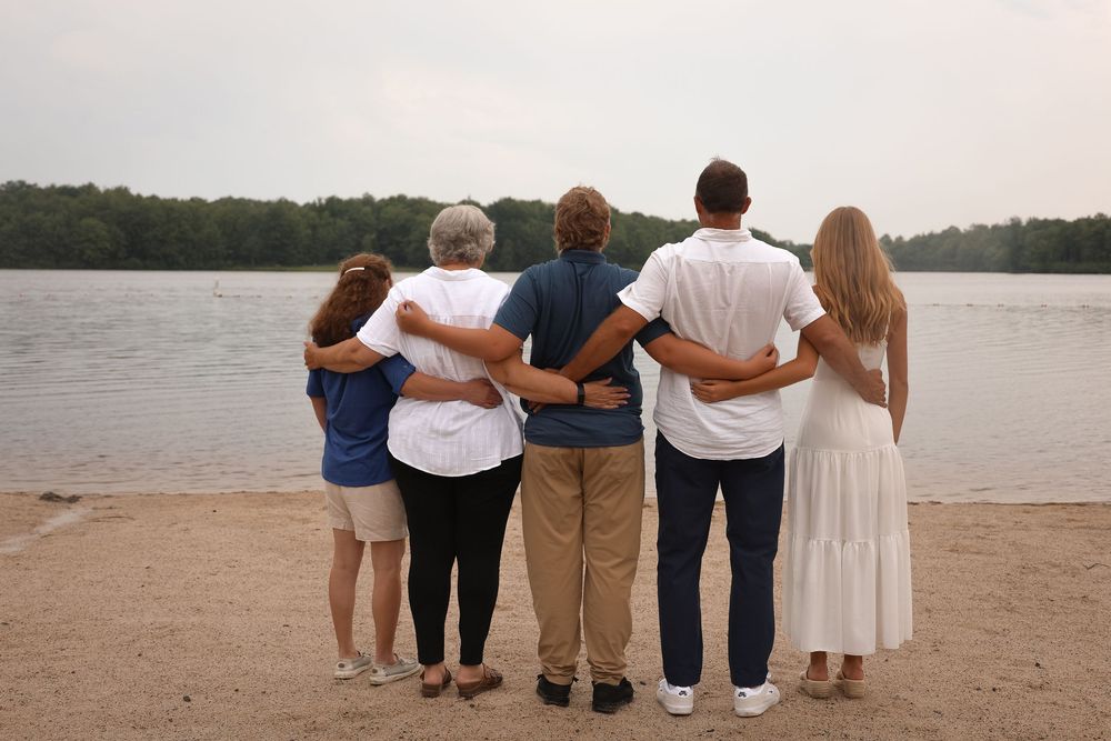 Five people stand together with their backs to camera, looking out over a peaceful lake from a sandy shore.