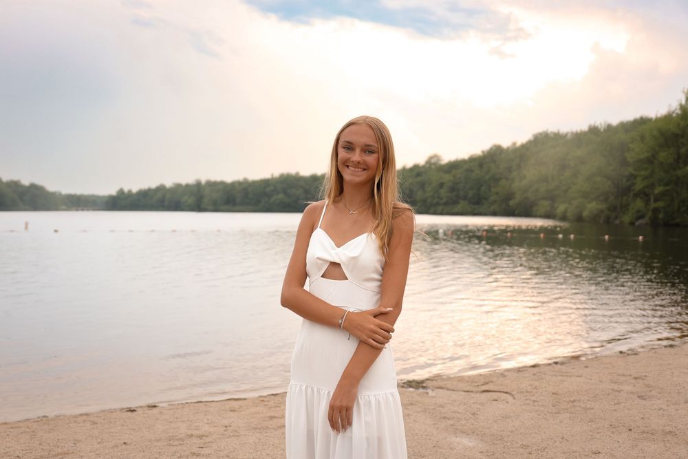 A person in white dress smiles near a lakeside beach during sunset while walking along the shoreline.