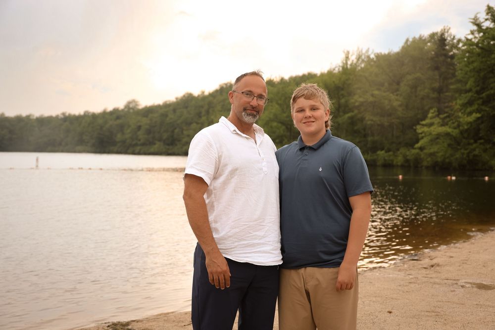Two people stand close together on a beach with a tranquil lake and wooded shoreline behind them.