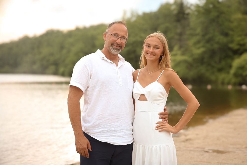 A couple poses lovingly together on a sandy beach during sunset with water and trees in the background.