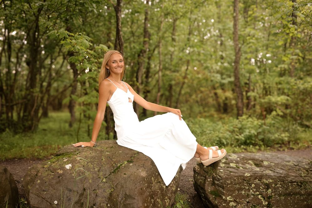 A person in a white dress sits elegantly on a large rock in a wooded forest setting.