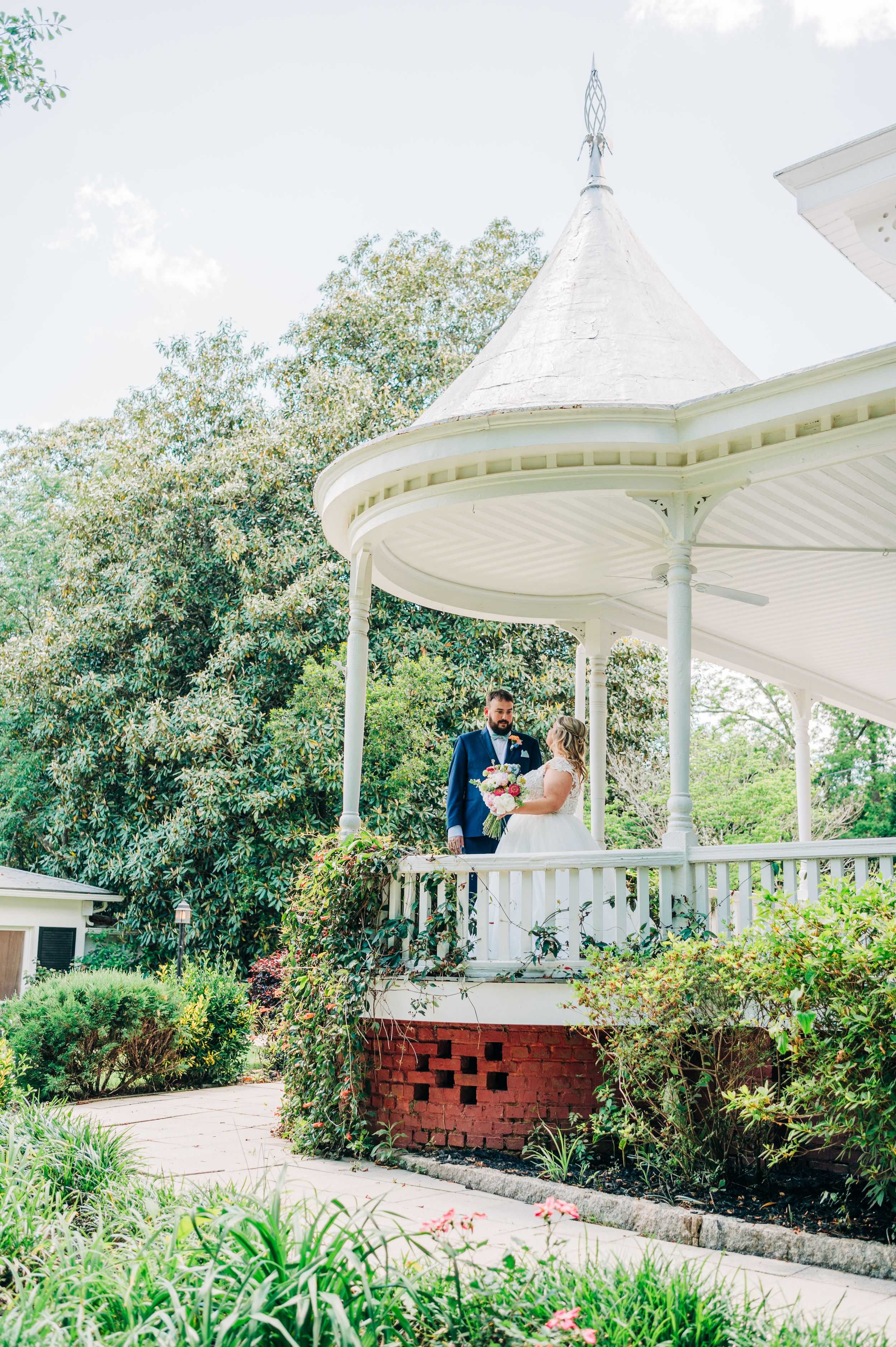 A white gazebo pavilion set in a lush garden landscape with brick accents and flowering plants along the pathway.