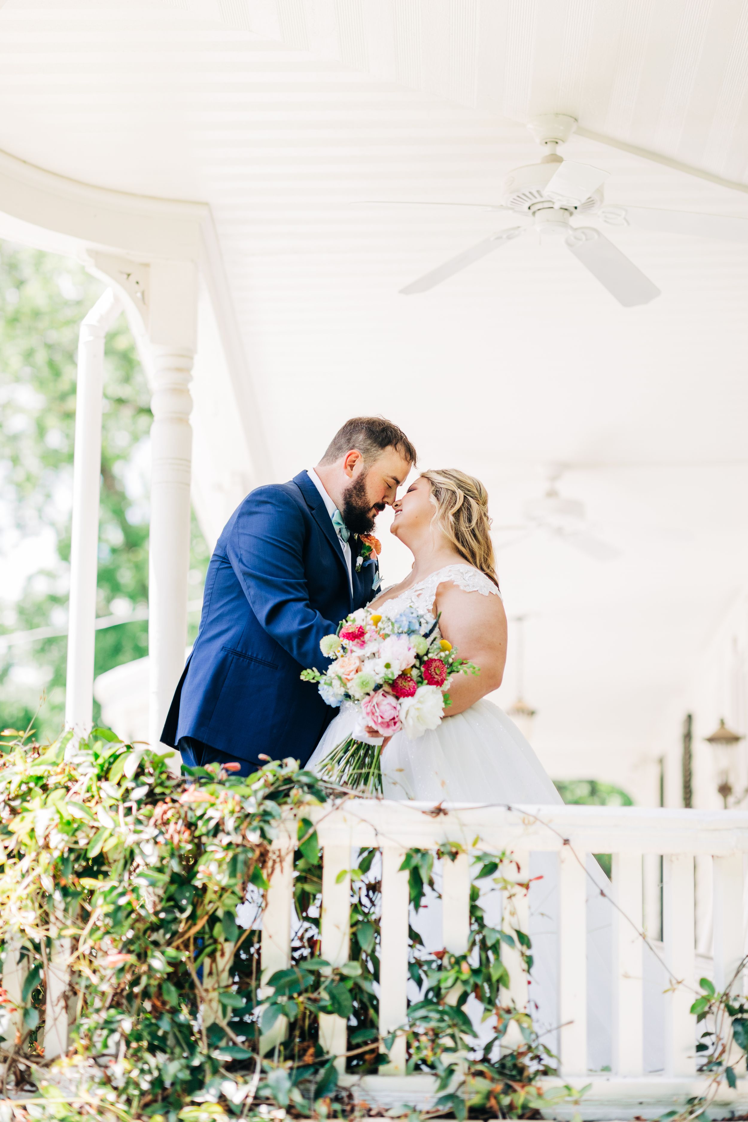 A romantic wedding moment shared on a white porch with lush greenery and beautiful floral bouquet.