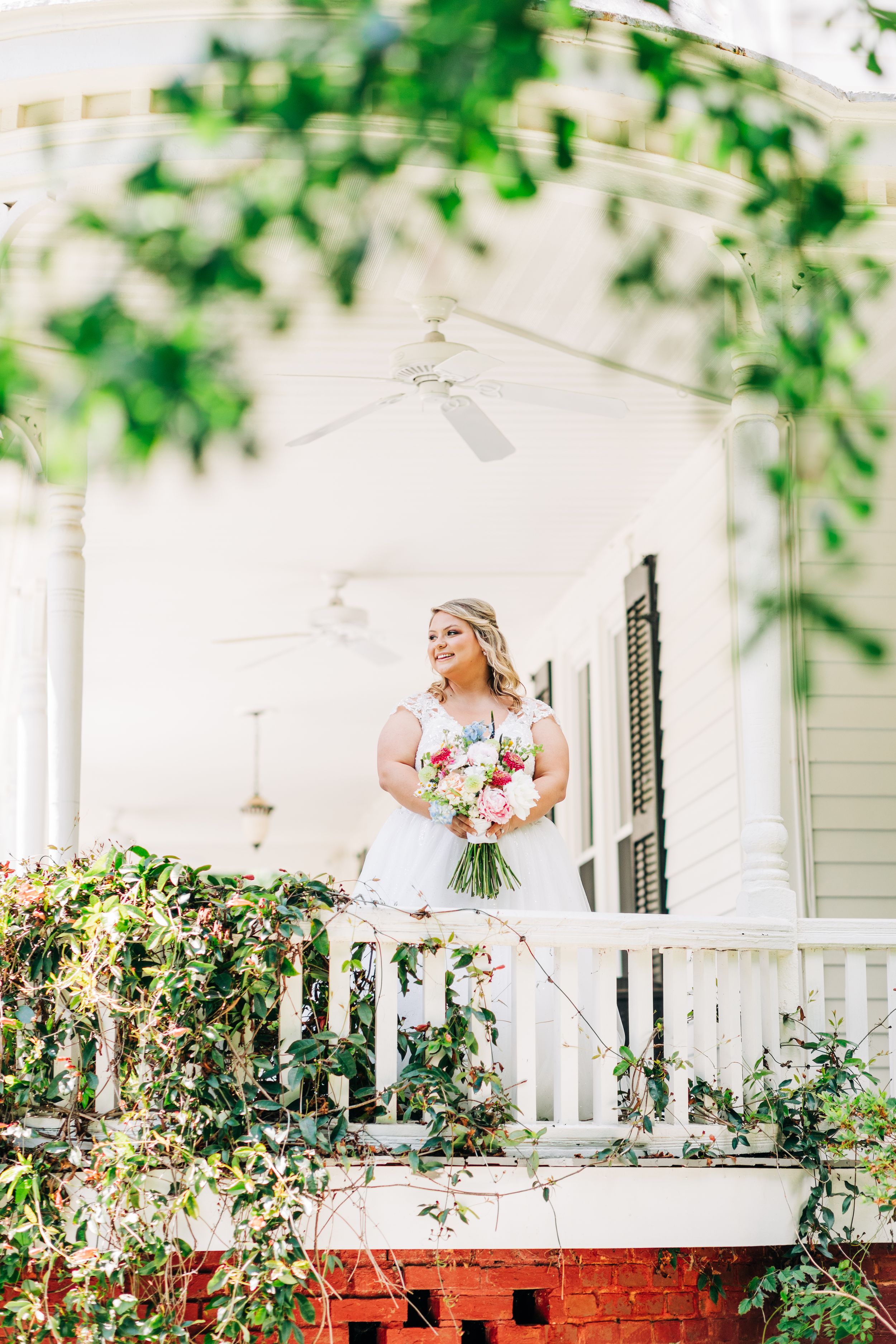 A bride poses on a white porch surrounded by lush greenery and red brick accents in a sunny garden setting.
