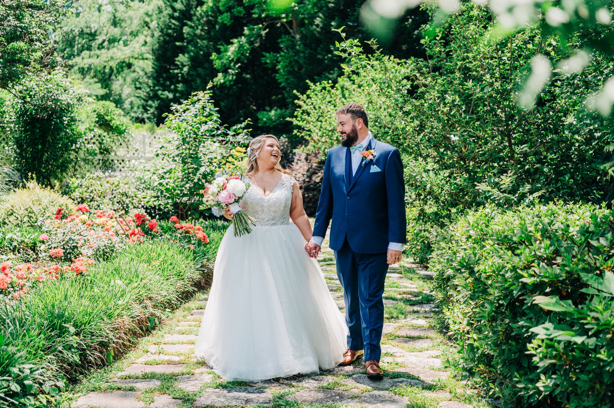 Two people in wedding attire walk together on a garden path surrounded by lush greenery and flowers on a sunny day.