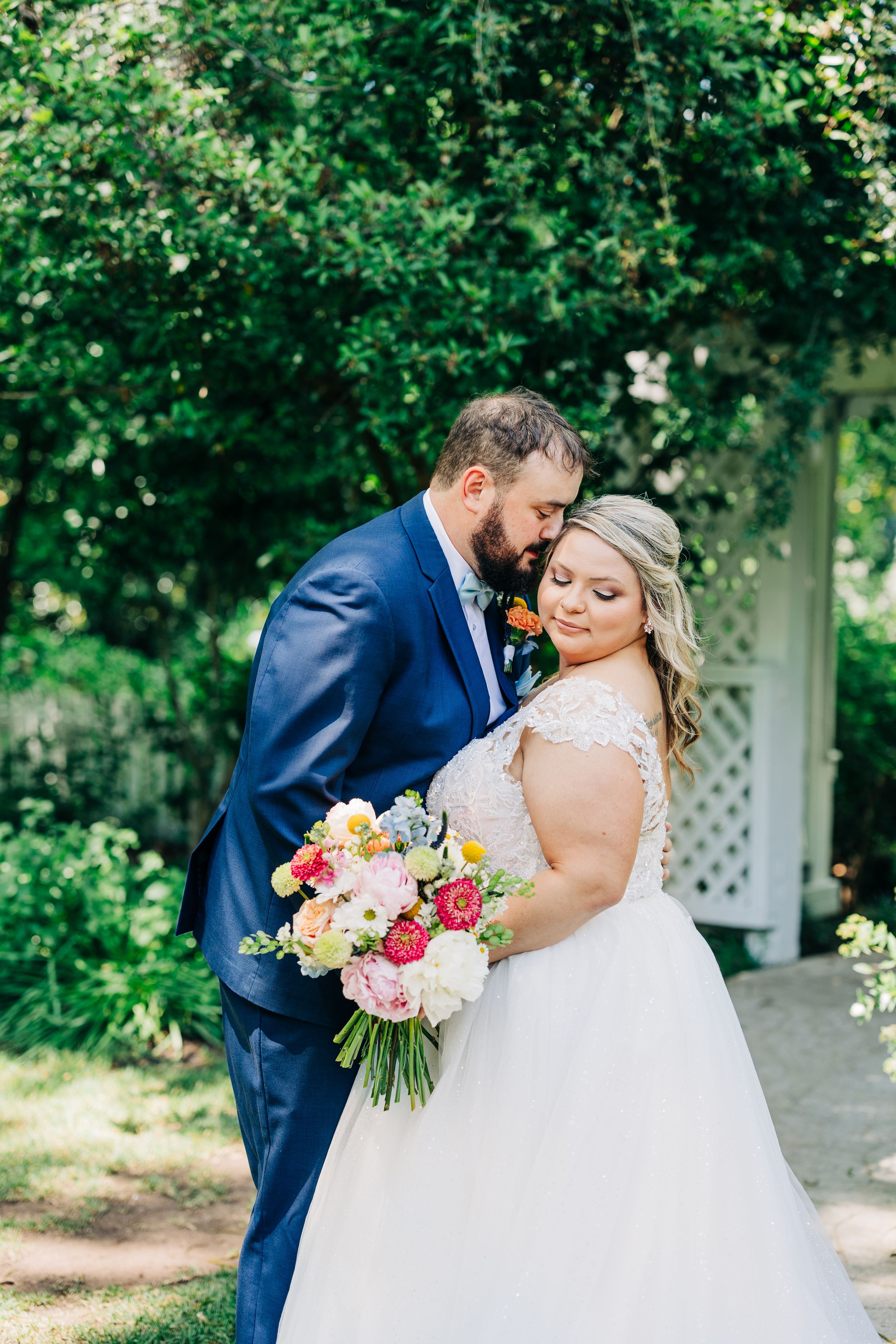 A romantic wedding couple shares intimate kisses in a garden with white lattice and lush greenery.