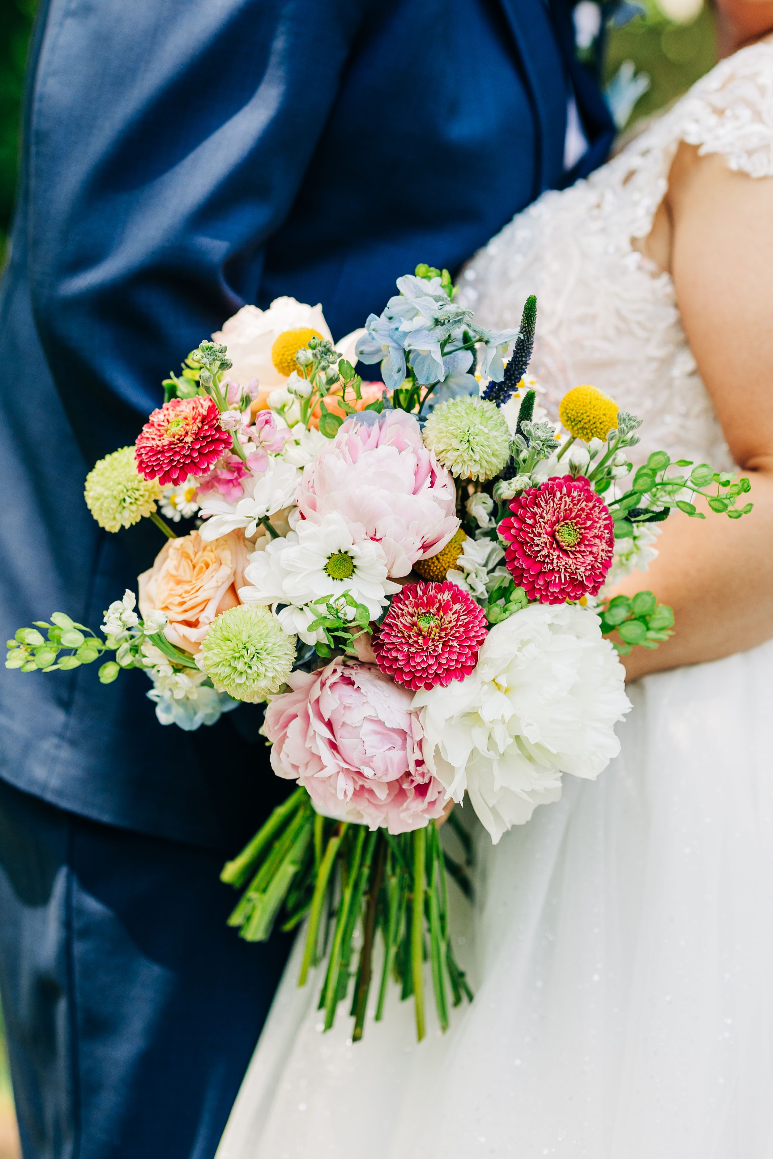 A wedding bouquet with pink peonies, white flowers, red dahlias and greenery against a navy blue background.