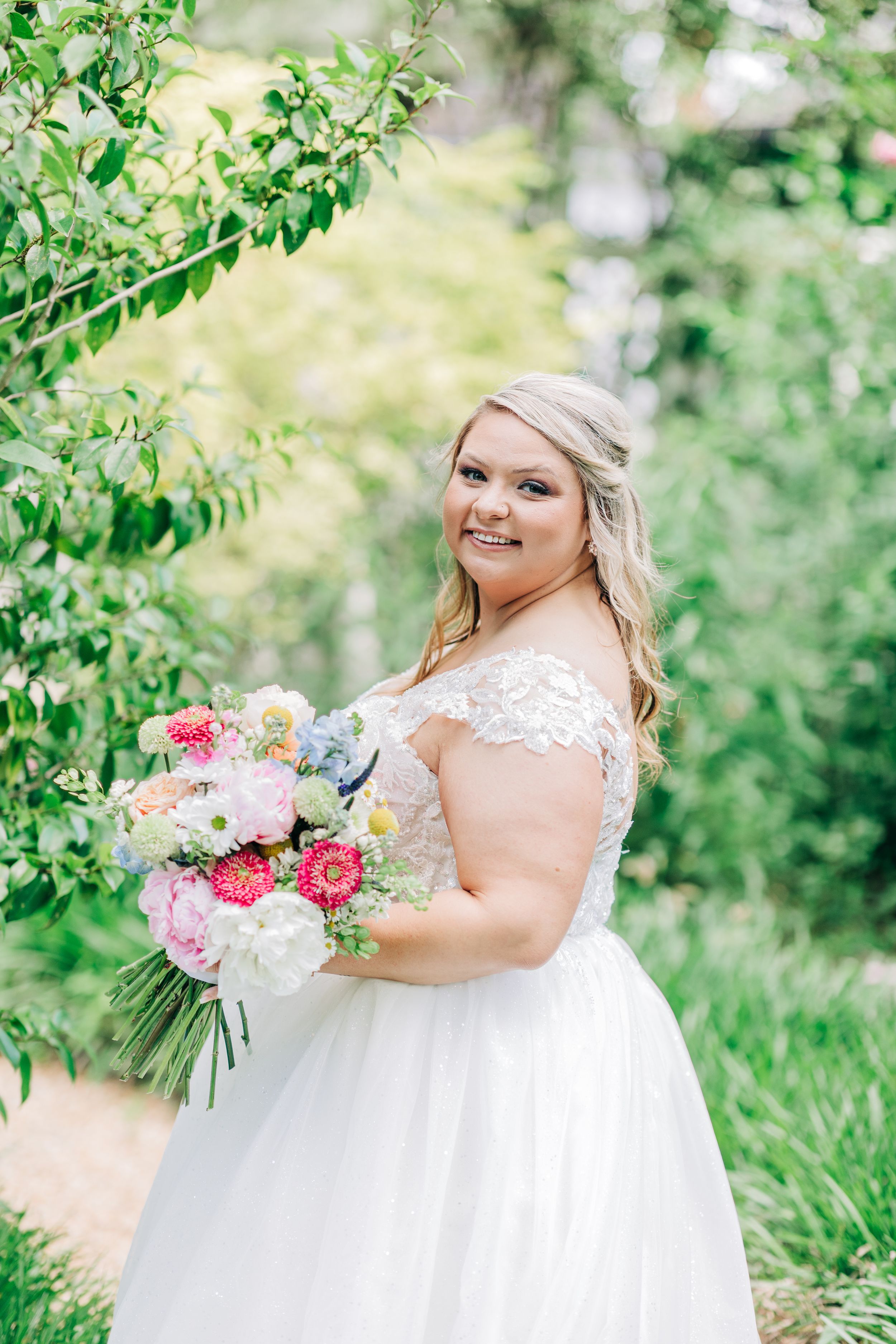Elegant bride in a flowing white dress holds a romantic pink and white bouquet against a lush garden backdrop.