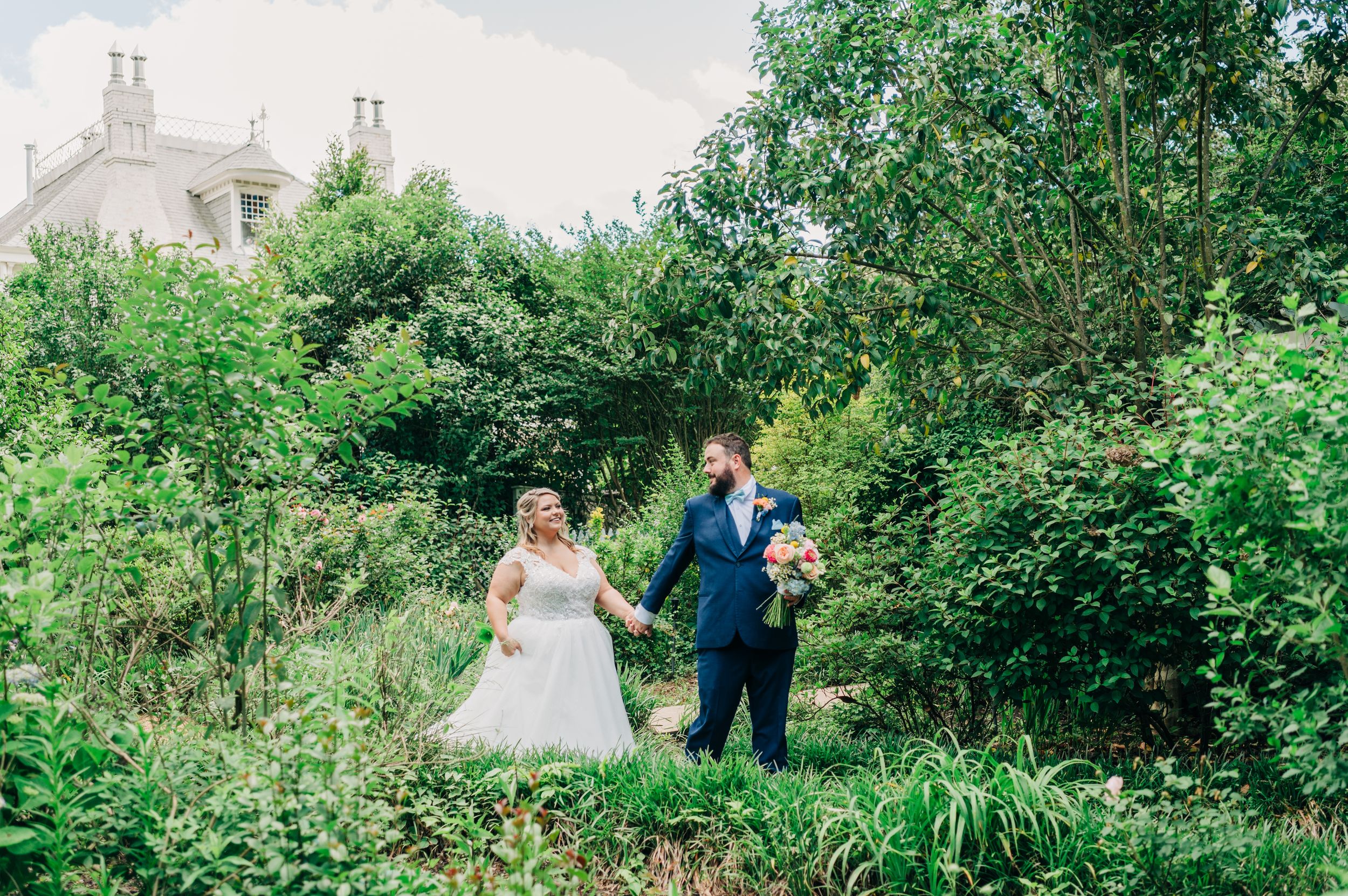 A romantic newlywed couple walks together through lush green gardens surrounded by flowering foliage.