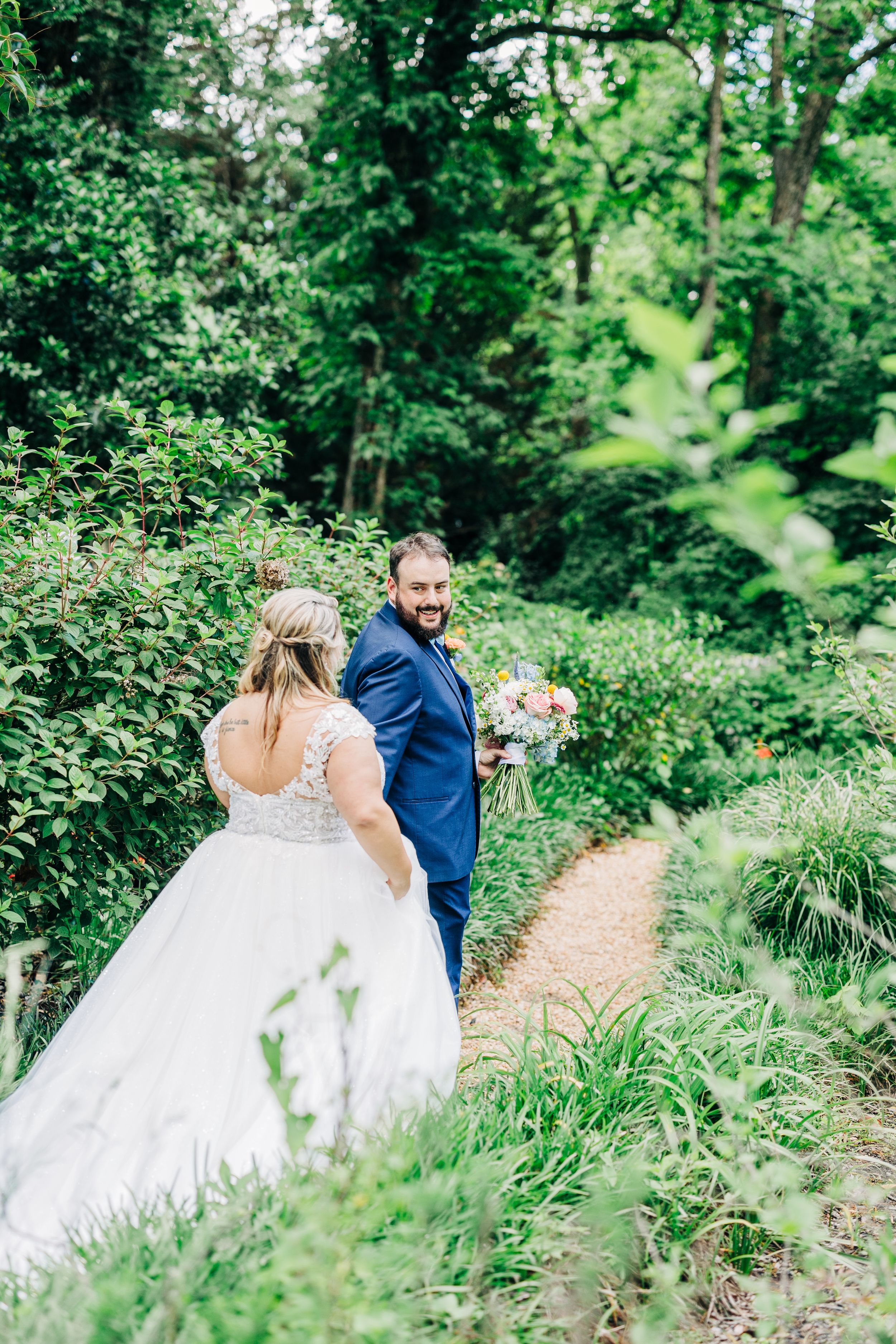 A couple walks along a lush garden path, with the bride in a white gown and groom in navy suit surrounded by greenery.