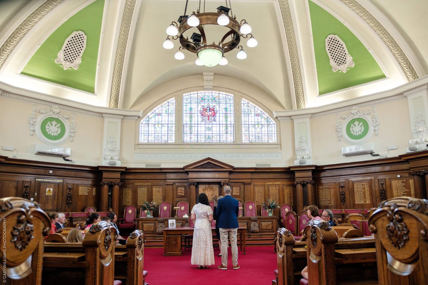 Historical courthouse interior with vaulted ceilings, ornate chandeliers, and pink carpet leading to elegant wooden indoor balconies.