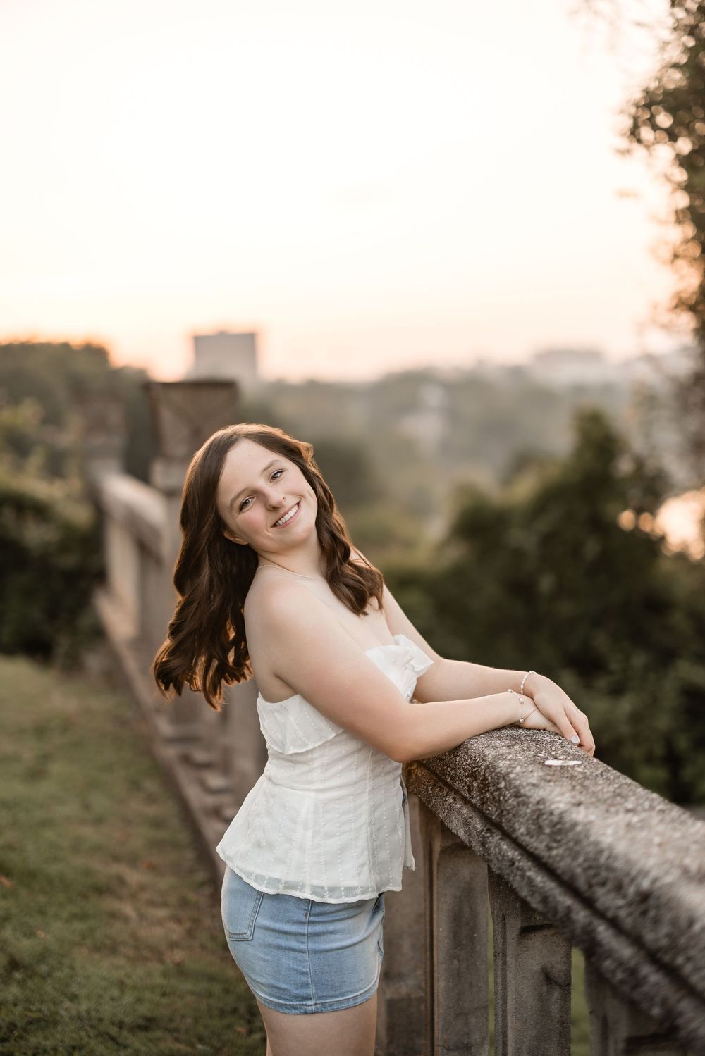 Young woman in white sleeveless top and denim shorts poses along wooden fence at sunset.