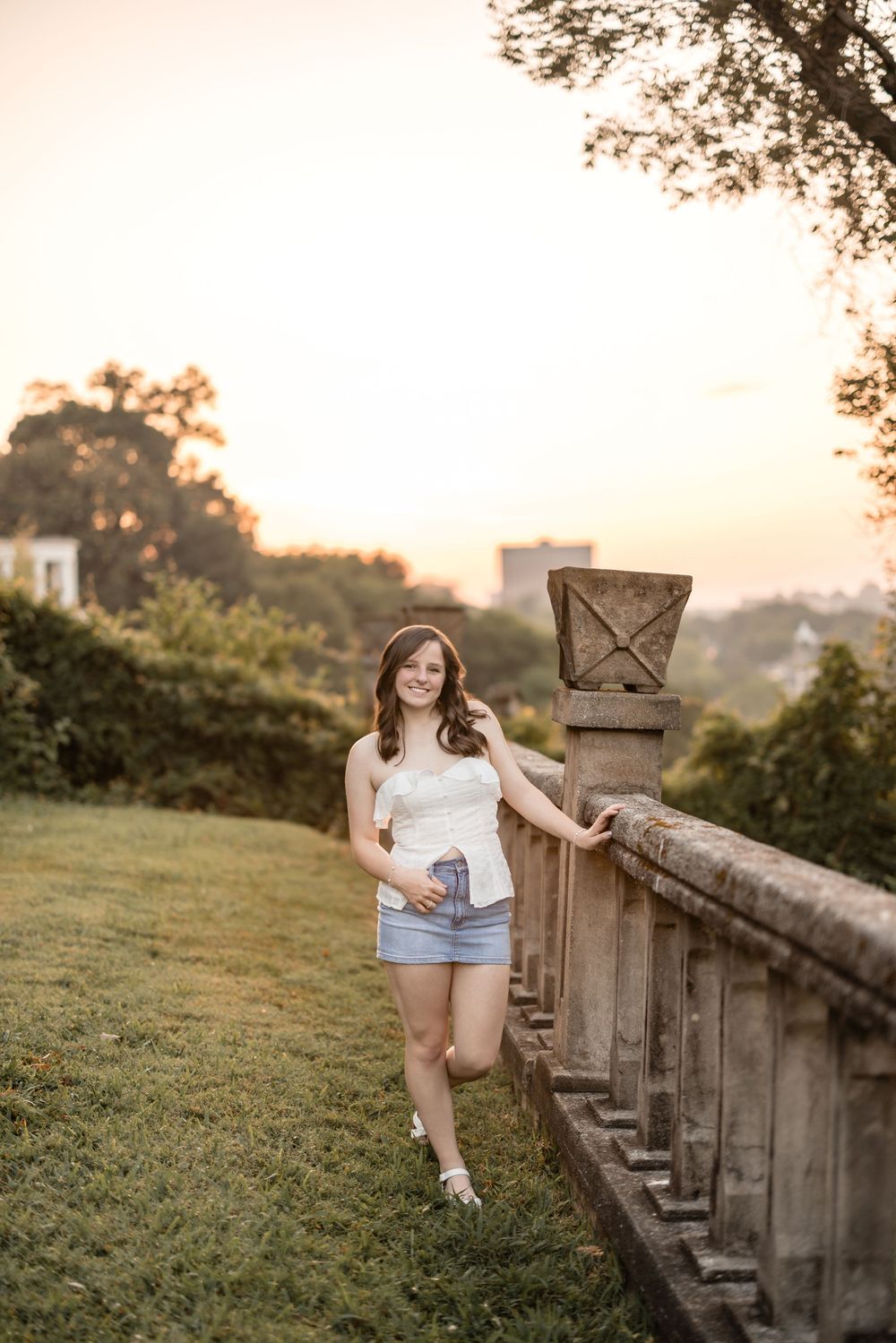Someone stands by a stone balustrade on a grassy hill during golden hour sunset with trees in the background.