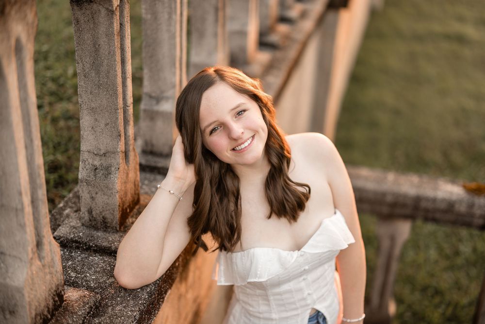 Woman in white dress poses outdoors in natural lighting with wooden post backgrounds.