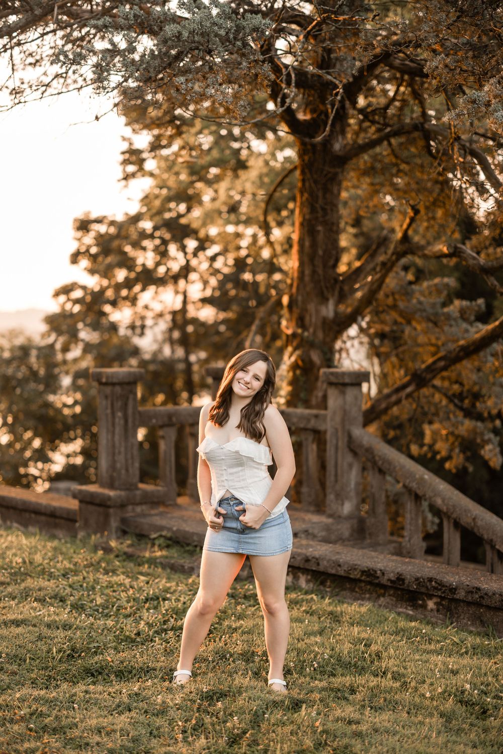 A photographer stands on a grassy hill during golden hour sunset with stone railing and autumn trees in background.