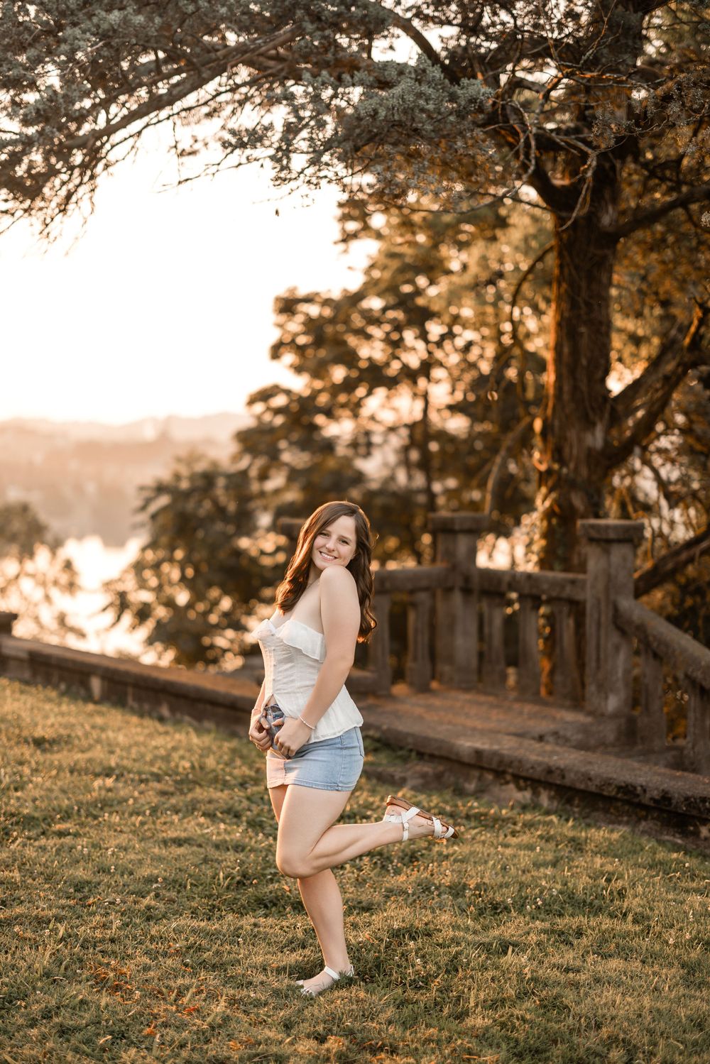 A woman poses outdoors at sunset on grassy hillside with golden light filtering through trees in background.