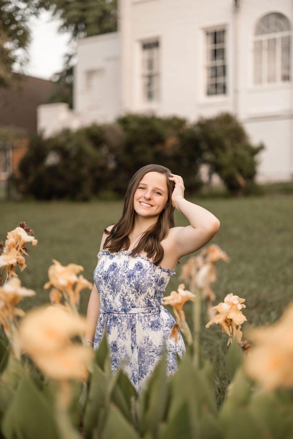 A person in a blue and white floral dress poses in a garden with blooming peach-colored irises and a white building behind.