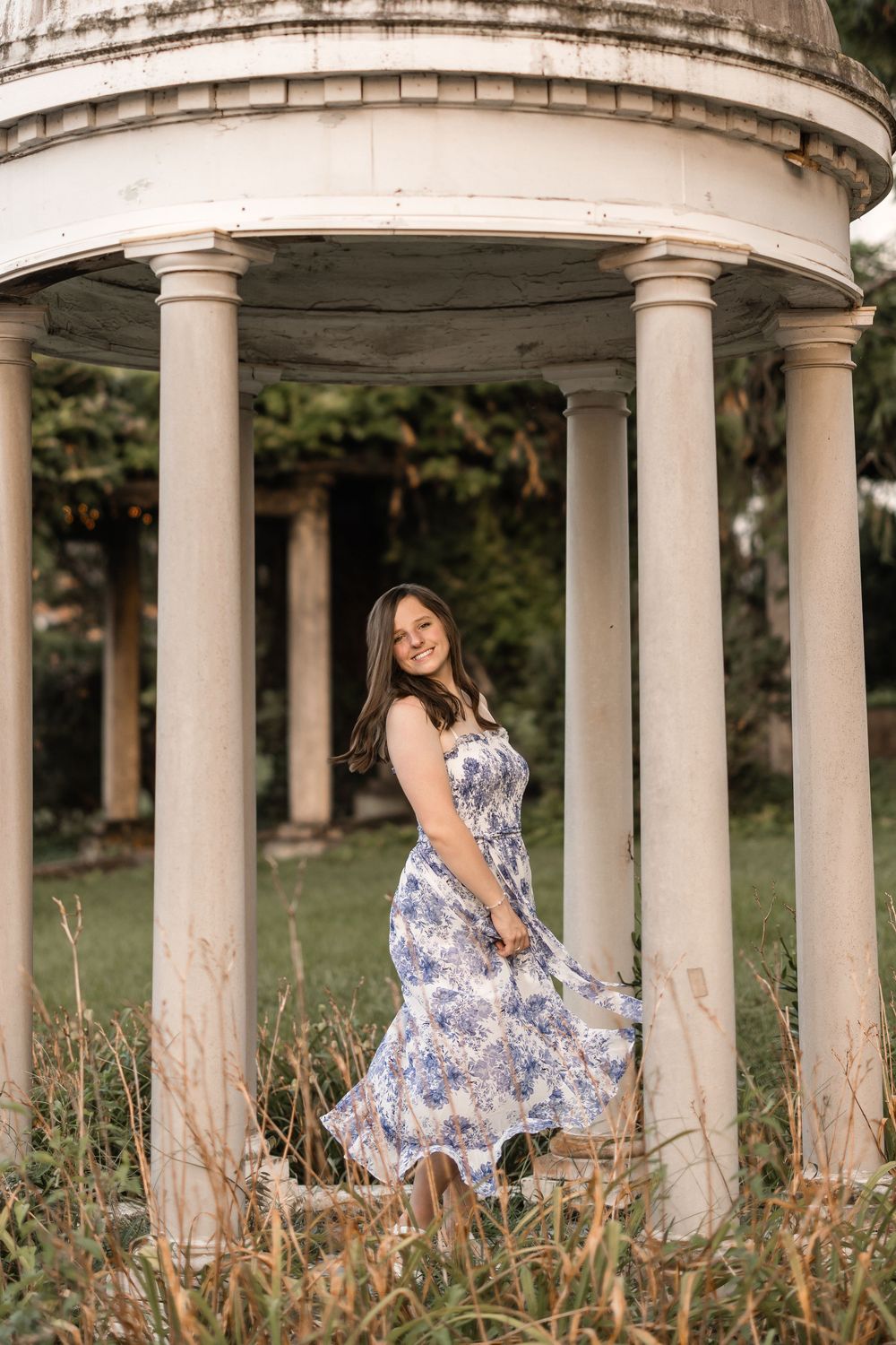 A blue and white floral dress flows gracefully among white stone columns in an outdoor garden setting.