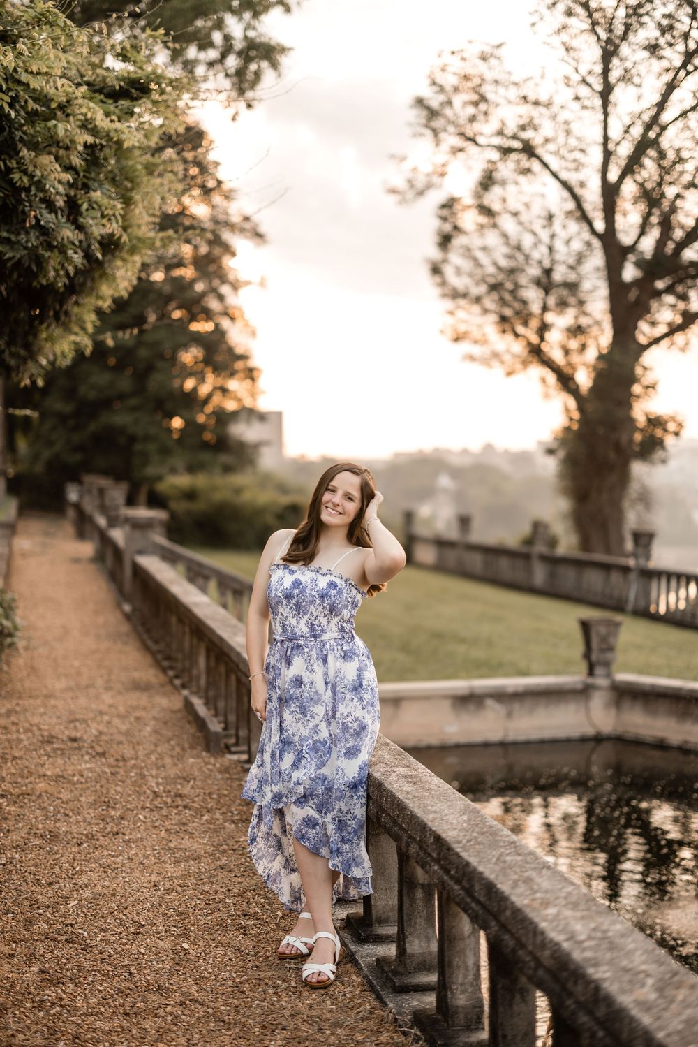 A figure in a floral blue and white summer dress walks along a gravel path by a stone wall during sunset.