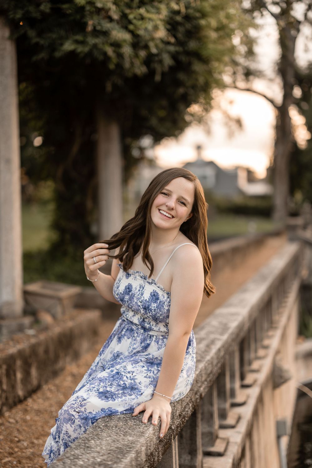 Woman in blue and white floral dress poses on stone wall during golden hour photo session.