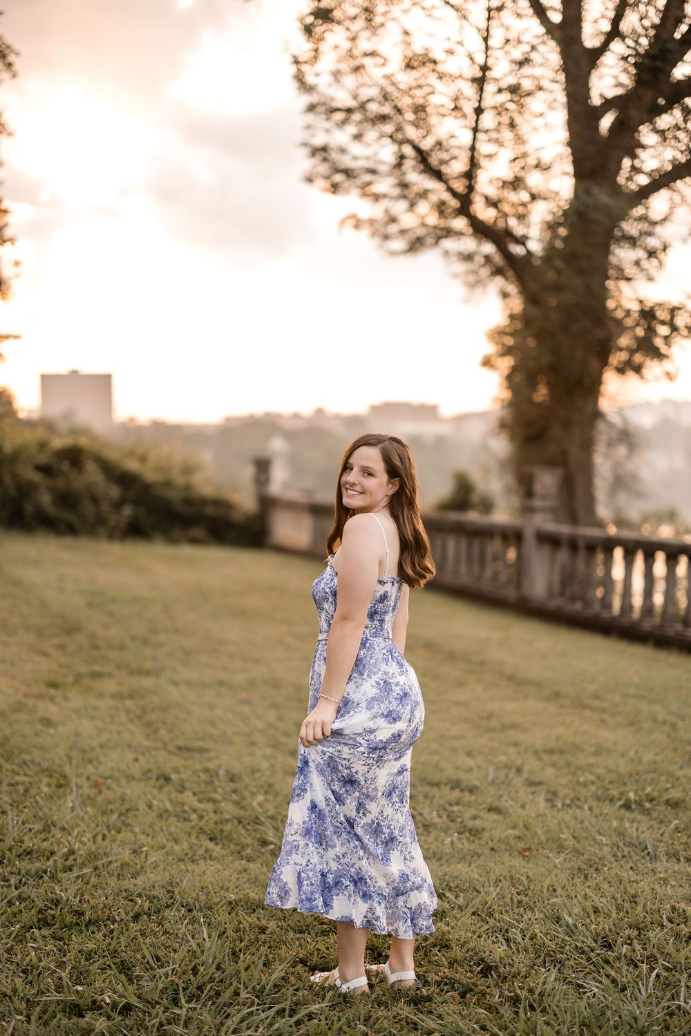 Woman in blue and white floral dress poses on grassy hill during golden sunset with city buildings in background.