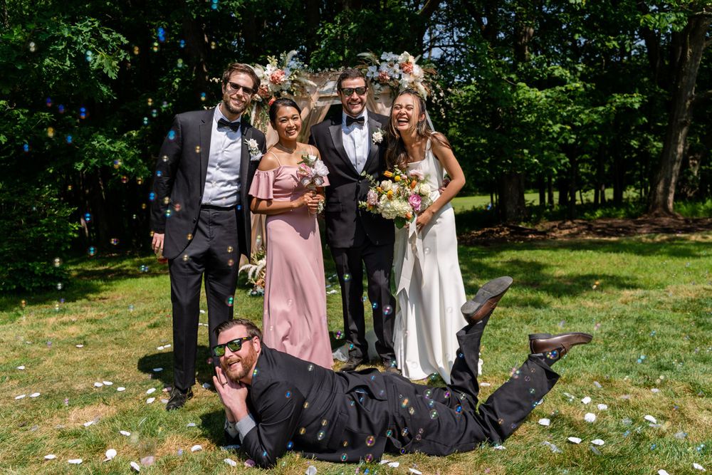 Wedding party poses playfully on grass with one person lying down in front.