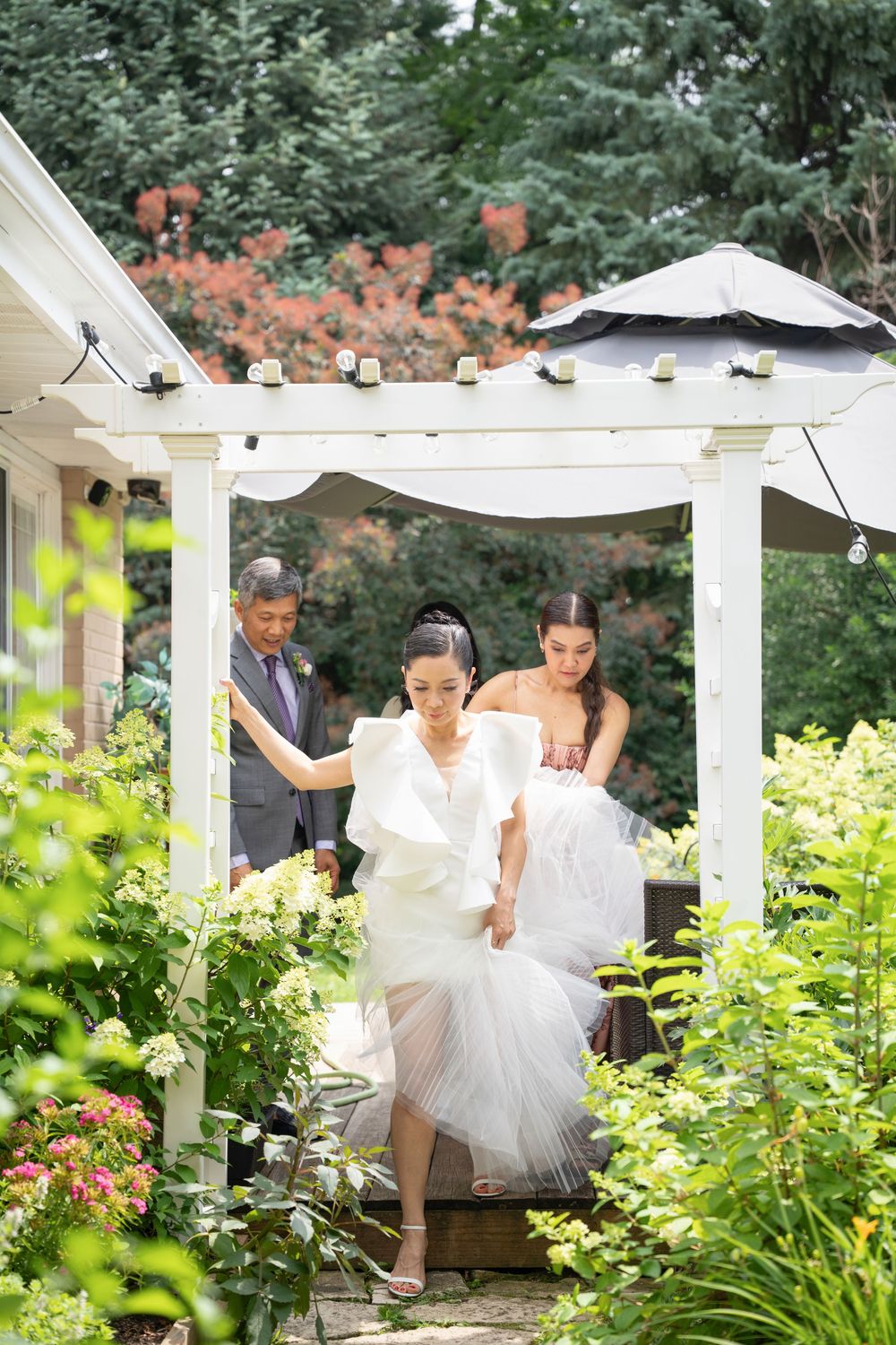 People wearing white dresses gather under an outdoor white pergola surrounded by lush greenery and flowers on a sunny day.