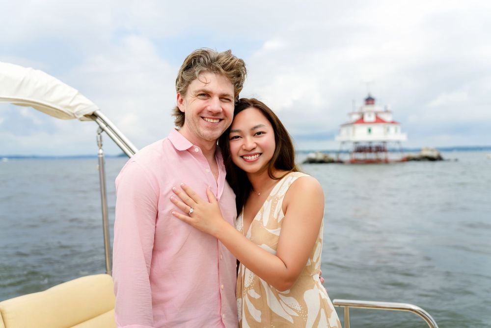 A couple smiles together during a boat outing on the water with a lighthouse and cloudy skies in the background.
