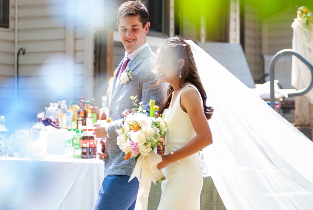 A couple in wedding attire standing together at an outdoor reception with sunlight streaming through.