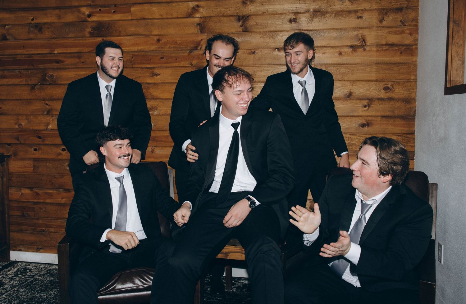 Groomsmen in black suits sharing laughs and conversations against a rustic wooden wall background during wedding festivities.