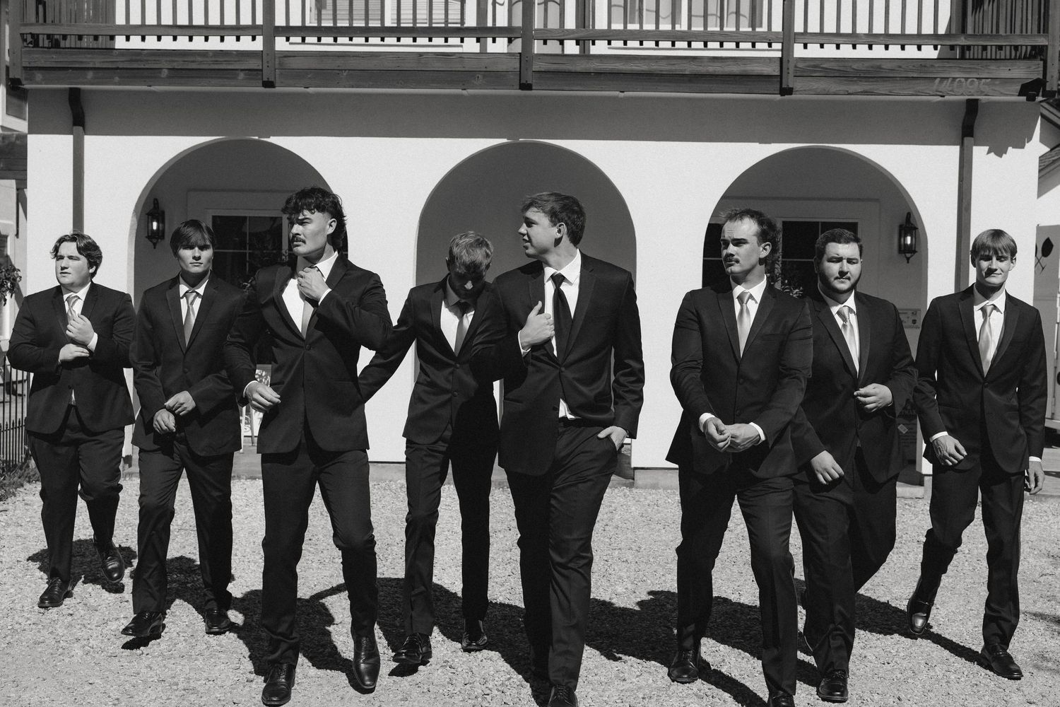 Groomsmen in classic black suits pose together outdoors in front of an arched colonnade in black and white photography.