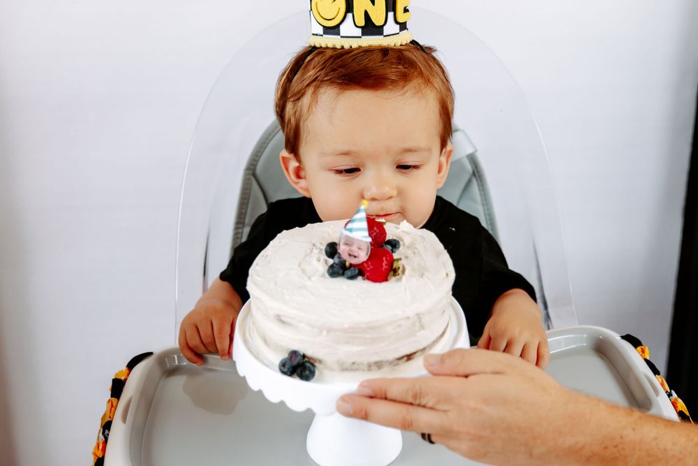 Small child in high chair with birthday crown looking down at white frosted cake.