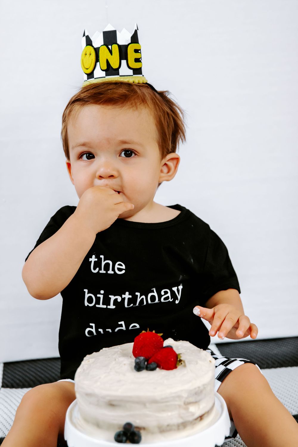 Small child wearing a birthday crown and black t-shirt tasting frosting from a white cake with berries.