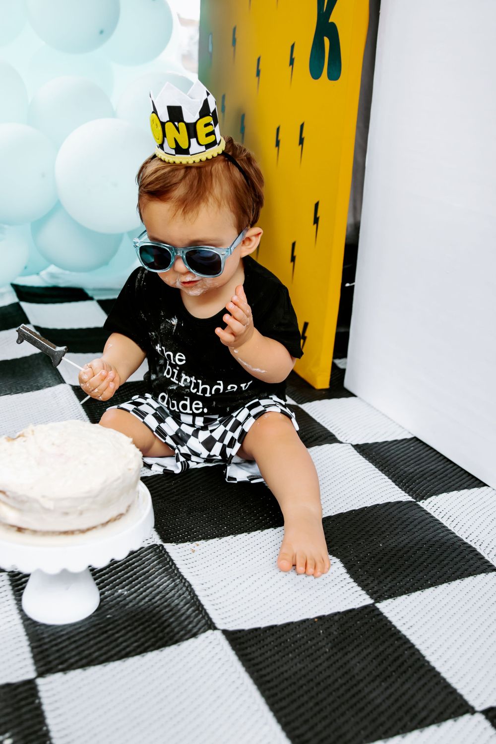 Small child in sunglasses and crown sitting on checkered floor with birthday cake.