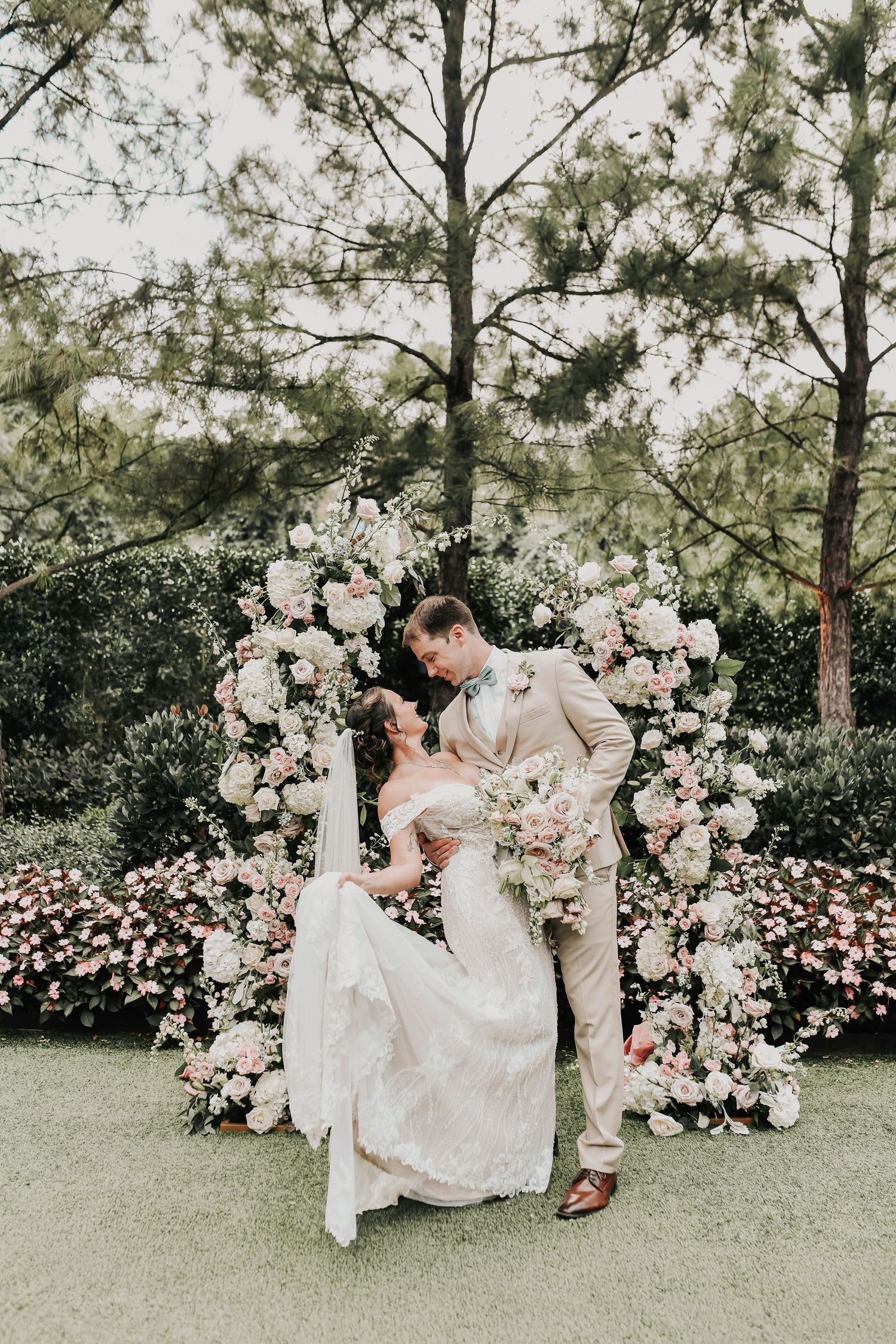 A groom in a beige suit dips his bride in front of their wedding altar covered in greenery and ivory florals